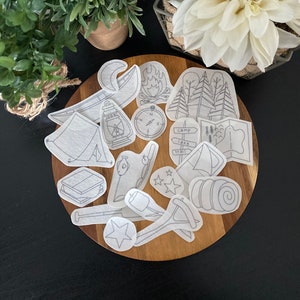 May include: A set of 16 white camping themed iron-on patches. The patches include a tent, campfire, compass, canoe, stars, a lantern, a map, a hiking stick, a marshmallow, a sleeping bag, and a sign that says "Camp, Lake, Trail".