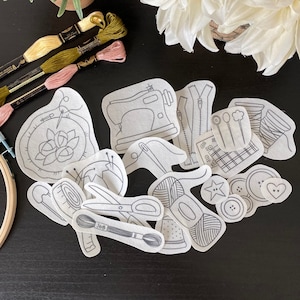 May include: A set of 14 white iron-on transfer designs for embroidery projects. The designs include a sewing machine, a zipper, a needle and thread, a spool of thread, a button, a heart, a flower, and other sewing-related items.