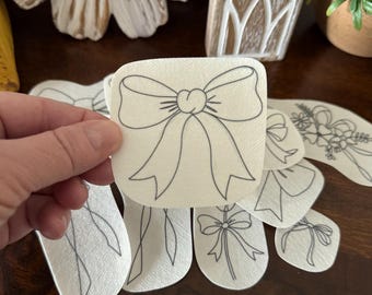 May include: A collection of white, die-cut bow-shaped embellishments with black outline details. The bows vary in style and size, with a variety of shapes. The embellishments are on a dark brown wooden surface, with decorative wooden objects in the background.