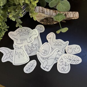 May include: A collection of dinosaur-themed fabric patches in various shapes and sizes. The patches feature black outlines of different dinosaurs, an erupting volcano, and other related designs. The patches are arranged on a dark surface.