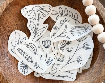 Op de afbeelding: Een verzameling witte bloemenstickers met zwarte omtrekken, met verschillende bloemen- en bladerontwerpen. De stickers liggen in een houten kom, omringd door houten kralen en een jute touw.