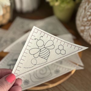 May include: A triangular banner with a bee illustration and honeycomb design. The bee is outlined in black, with a dotted flight path. The banner has a ruler-like border and is likely part of a craft or DIY project.