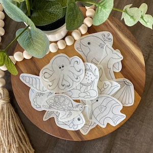 May include: A set of 10 white felt ocean animal cutouts, including a whale, octopus, turtle, fish, and seashells. The cutouts are on a wooden surface.