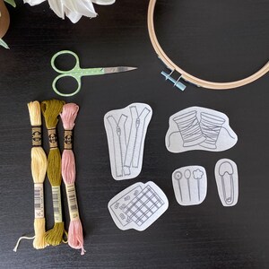 Op de afbeelding: Een set borduurbenodigdheden, waaronder drie spoelen borduurgaren in groen, geel en roze, een paar groene borduurscharen, een ring en vijf borduurpatronen. De patronen omvatten een rits, een spoel garen, een knoop, een veiligheidsspeld en een raster.