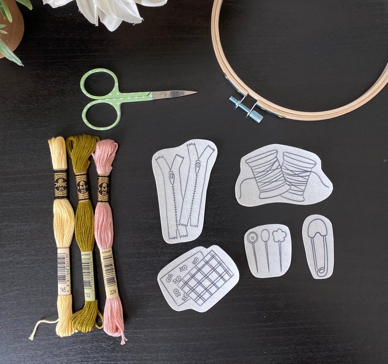 Op de afbeelding: Een set borduurbenodigdheden, waaronder drie spoelen borduurgaren in groen, geel en roze, een paar groene borduurscharen, een ring en vijf borduurpatronen. De patronen omvatten een rits, een spoel garen, een knoop, een veiligheidsspeld en een raster.
