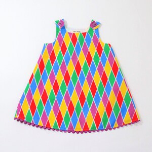 May include: A sleeveless dress featuring a vibrant harlequin diamond pattern in red, yellow, green, blue, and purple. The garment has two purple buttons at the shoulders and a purple scalloped trim along the bottom hemline with a white label.