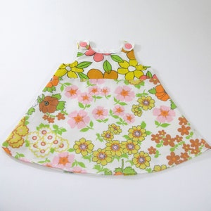 May include: A sleeveless A-line dress featuring a white base with a vibrant floral pattern in shades of yellow, orange, pink, and green. The garment includes two round pink buttons at the shoulder straps and a flared silhouette with a hemline.