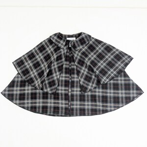 May include: A black, white and red plaid cape featuring a rounded collar and a single button closure at the neckline. The garment has a layered design with a shorter capelet overlaying a longer, flared base, creating a structured silhouette.