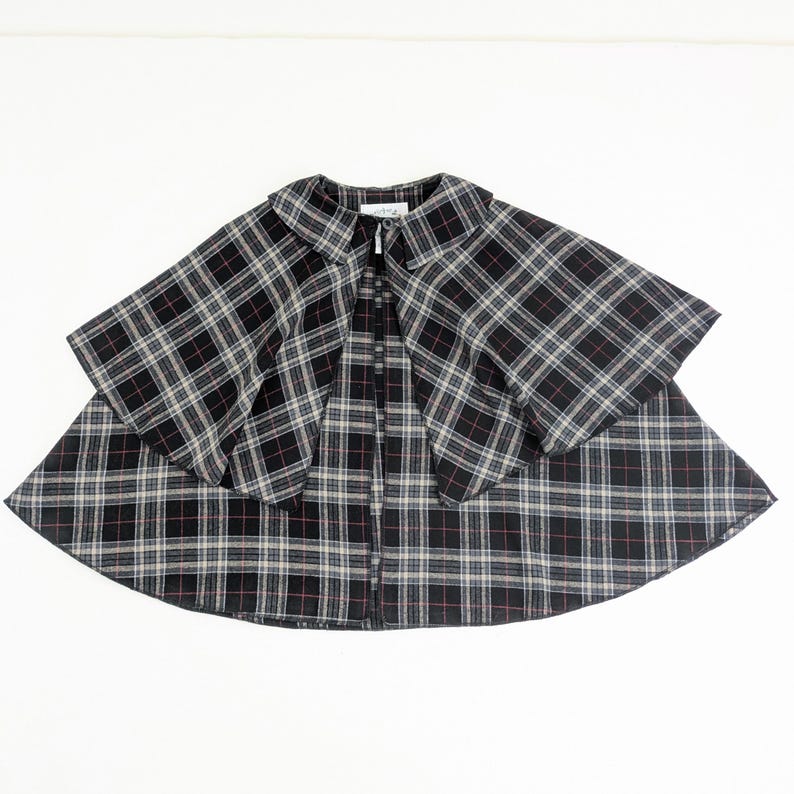 May include: A black, white and red plaid cape featuring a rounded collar and a single button closure at the neckline. The garment has a layered design with a shorter capelet overlaying a longer, flared base, creating a structured silhouette.