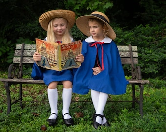 May include: Two blue capes with white collars and red ribbon ties, worn with straw hats, white knee-high socks, and black shoes. One cape features a book titled Madeline's Rescue by Ludwig Bemelmans, depicting a building and trees in an illustration.