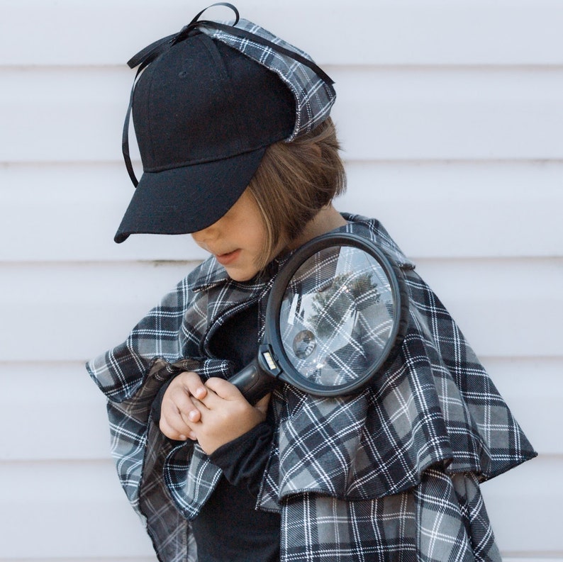 May include: A black baseball cap with a matching plaid cape featuring a white and grey check pattern. The ensemble includes a black magnifying glass with a circular lens. The cape has a wide collar and drapes over the shoulders in a layered style.