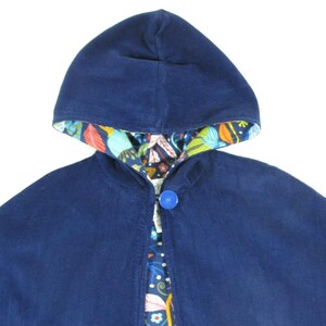 May include: A solid navy blue hooded cape featuring a single large blue button closure at the neck. The interior lining of the hood and the front opening displays a colourful botanical pattern with leaves and flowers in orange, teal, and white.