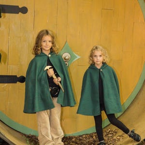 May include: Two forest green hooded capes with a single metallic button closure at the neck. The capes are worn over black shirts, with one cape paired with tan trousers and the other with black leggings. One cape features a gold-toned toy sword.