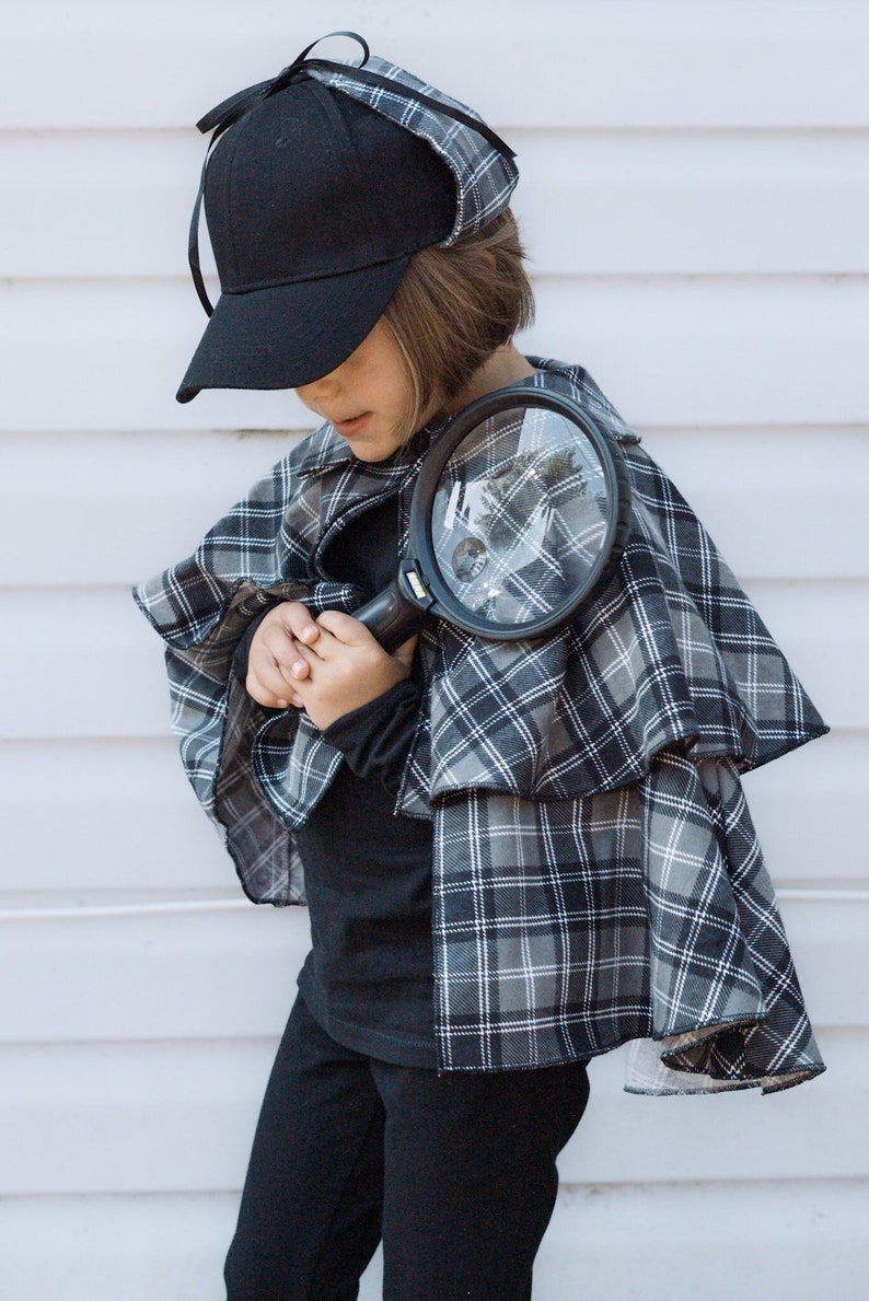 May include: A black baseball cap with side flaps and a black ribbon tie, paired with a grey and black plaid cape featuring a collar and layered fabric. A black magnifying glass with a circular lens is held in front of the plaid cape garment.