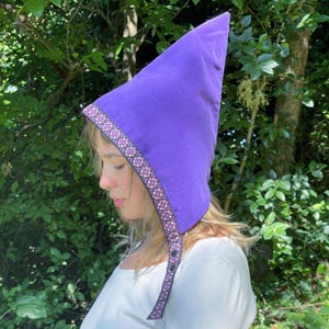May include: A vibrant purple hood with a pointed top. The hood features a decorative trim with a repeating floral pattern in pink, white, and black. The hood has a strap with buttons for closure. The background is a natural outdoor setting.