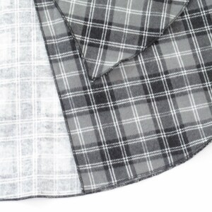 May include: A circular fabric piece featuring a grey, black, and white plaid pattern. The textile displays a darker, saturated tartan design on the top layer and a lighter, faded version of the same checkered print on the underside, with a finished edge.