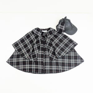May include: A black, white and red plaid cape with a wide collar and a front button fastening, accompanied by a matching dark grey baseball cap featuring a plaid fabric accent attached to the side, creating a coordinated set of outerwear accessories.