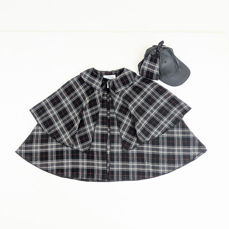 May include: A black, white and red plaid cape with a wide collar and a front button fastening, accompanied by a matching dark grey baseball cap featuring a plaid fabric accent attached to the side, creating a coordinated set of outerwear accessories.