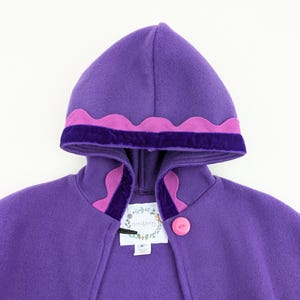 May include: A purple hooded cape with a scalloped pink trim and a dark purple velvet band. The cape features a pink button closure and a tag with floral illustrations.
