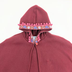 May include: A burgundy hooded cape with a decorative trim featuring pink pom-poms and a floral pattern in blue, pink, and brown. The cape has a single red button closure at the neck.