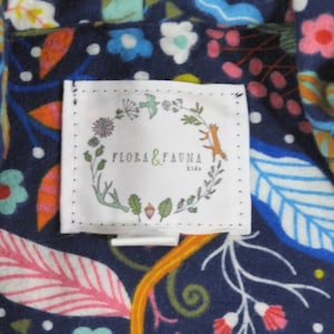 May include: A white rectangular clothing label with the text Flora & Fauna kids centred inside a circular wreath illustration featuring small leaves, flowers, a bird, and a fox, stitched onto a dark blue fabric with a colourful floral pattern.
