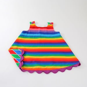 May include: A sleeveless dress featuring horizontal rainbow stripes in red, orange, yellow, green, blue, and purple. The garment has two purple buttons at the shoulders and a scalloped purple hemline that reveals a colorful geometric diamond pattern.
