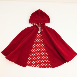 May include: A red hooded cape with a heart-shaped button closure. The cape's interior lining features a white heart pattern on a red background. The cape is made of a soft, velvety material, perfect for costumes or imaginative play.