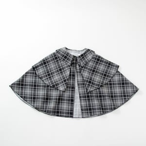 May include: A grey, black and white plaid cape featuring a wide, folded collar and a layered design. The garment has a flared, circular silhouette that opens at the front, showcasing a consistent tartan pattern throughout the fabric construction.