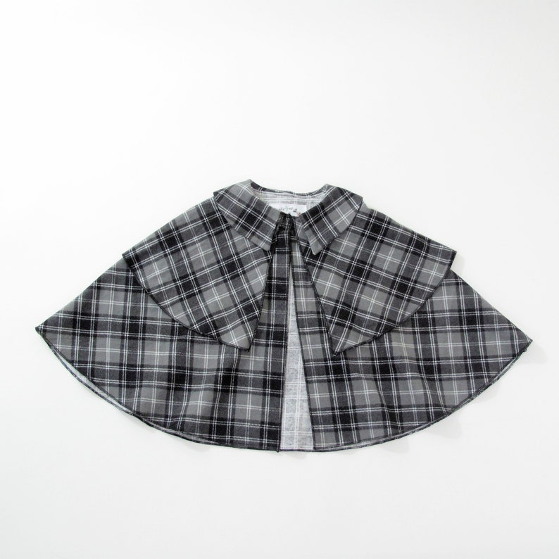 May include: A grey, black and white plaid cape featuring a wide, folded collar and a layered design. The garment has a flared, circular silhouette that opens at the front, showcasing a consistent tartan pattern throughout the fabric construction.