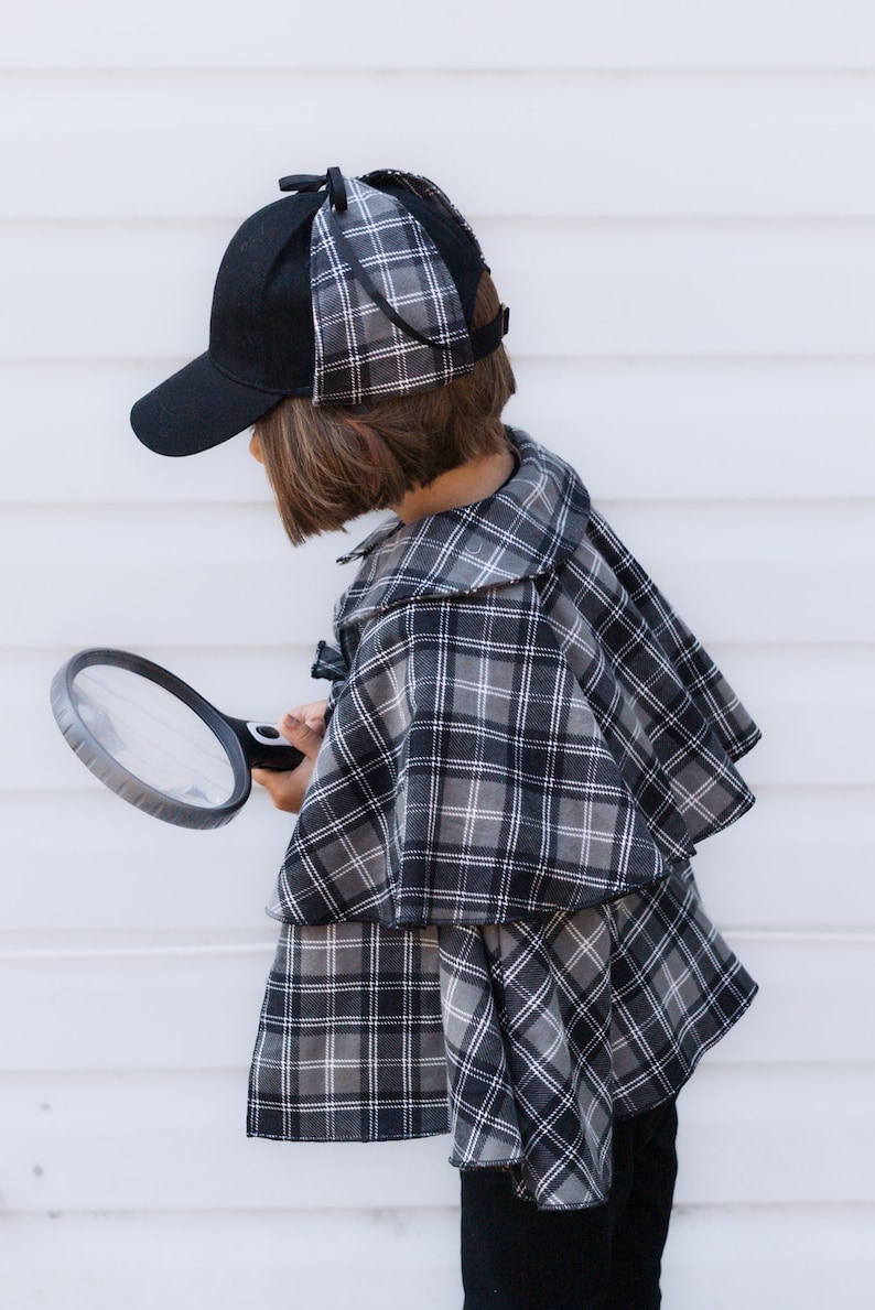 May include: A black baseball cap with plaid side flaps and a matching plaid cape featuring a grey, black and white tartan pattern. The ensemble includes a handheld magnifying glass with a black frame and a clear lens, completing the detective look.