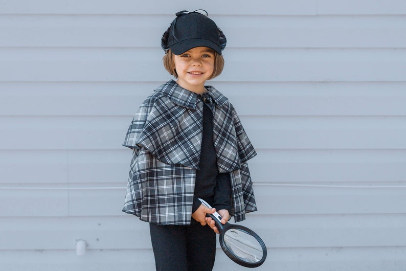 May include: A black deerstalker hat, a black long-sleeved shirt, black trousers, and a grey and black plaid cape with a collar. A black magnifying glass is held in the hand. The outfit is designed for a detective costume, featuring a classic pattern.