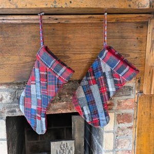 Può includere: Due calze di Natale con un design patchwork a quadri rossi, blu e grigi appese a un caminetto in legno. Le calze hanno un risvolto rosso e sono sospese da nastri colorati a fantasia.