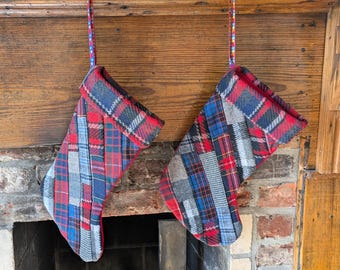 Pode incluir: Duas meias de Natal com um design xadrez de patchwork em vermelho, azul e cinza penduradas em uma lareira de madeira. As meias têm uma bainha vermelha e são suspensas por fitas coloridas e estampadas.
