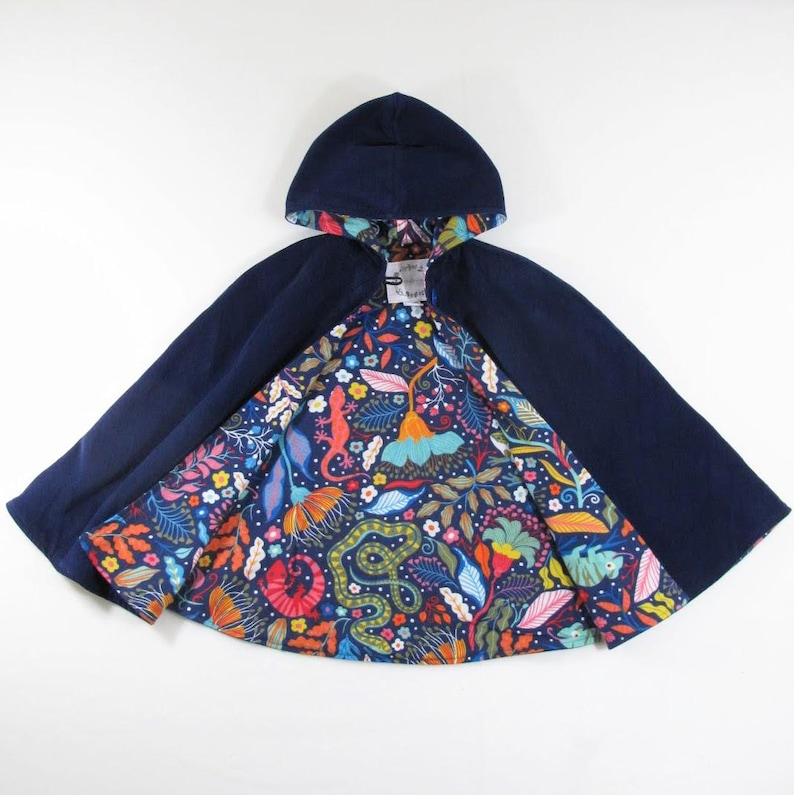 May include: A hooded cape featuring a solid dark navy blue exterior and a vibrant interior lining. The lining displays a colourful botanical pattern with illustrations of lizards, snakes, and various tropical flowers in shades of red, orange, yellow, and blue.