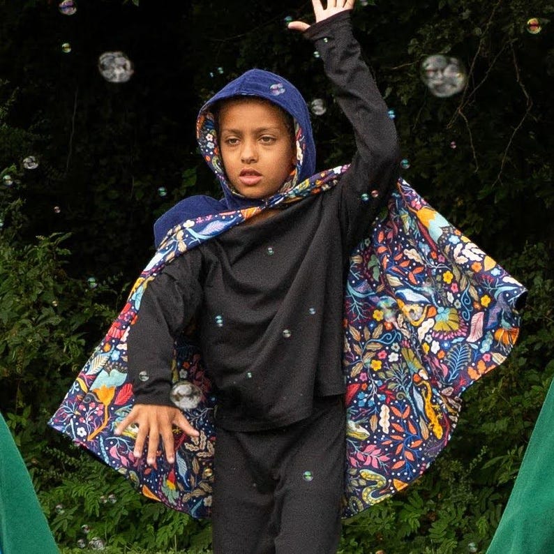 May include: A hooded cape featuring a dark blue base with a vibrant, multicoloured floral and botanical pattern. The cape is worn over a solid black long-sleeved shirt and black trousers, with the hood pulled up and the fabric draped over the shoulders.