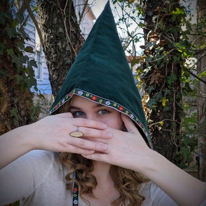 May include: A woman wearing a green hooded cloak with a floral trim. The hood is pulled over her head, and she is covering her face with her hands. She is wearing a ring on her finger.