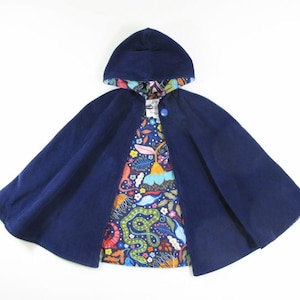 May include: A hooded cape in solid navy blue with a single blue button closure at the neck. The interior lining features a vibrant, multi-coloured botanical print on a dark blue background, showcasing stylised snakes, flowers, and leaves.