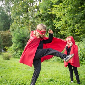 May include: A bright red hooded cape paired with a matching red eye mask, worn over a black long-sleeved shirt and black pants. The cape features a hood and drapes over the shoulders, creating a superhero-inspired costume set for active play.