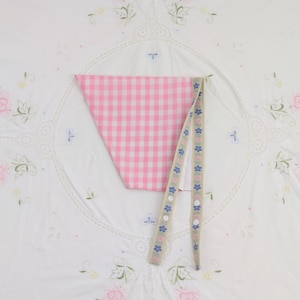 May include: A pink and white gingham fabric triangle with a beige ribbon featuring blue and pink floral designs and white buttons. The items are displayed on a white embroidered cloth with floral and lace details.