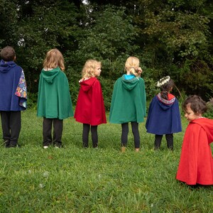 May include: A collection of hooded capes in solid colours including navy blue, forest green, and bright red. The capes feature attached hoods and vary in length, with some displaying patterned fabric linings visible along the edges and hood interiors.