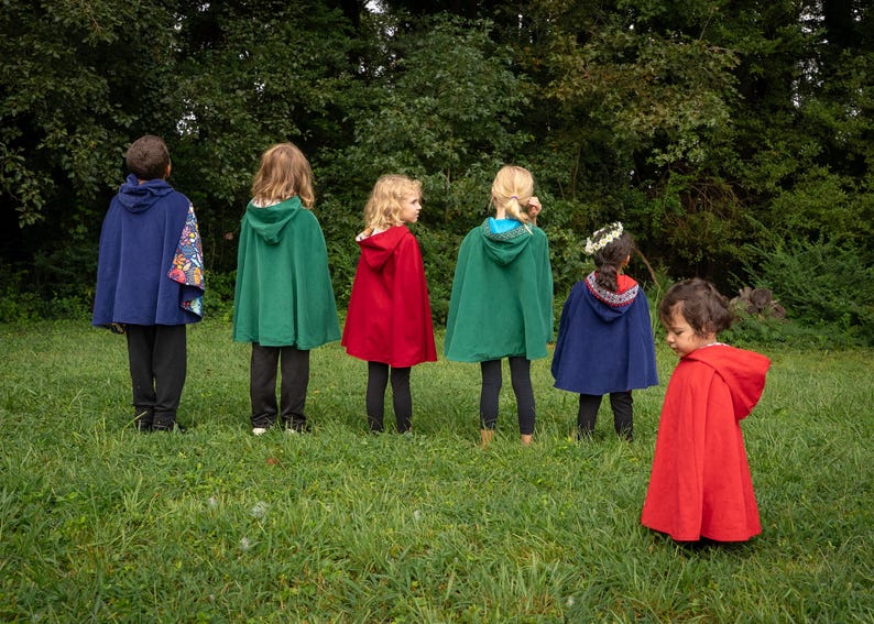 May include: A collection of hooded capes in solid colours including navy blue, forest green, and bright red. The capes feature attached hoods and vary in length, with some displaying patterned fabric linings visible along the edges and hood interiors.