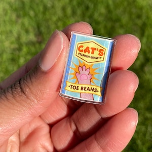May include: A rectangular acrylic pin featuring a cartoon design. The pin has a blue background with the words "CAT'S PREMIUM QUALITY" and "TOE BEANS." A pink cat paw is in the center, with yellow starbursts around it.