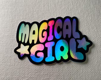 May include: A holographic sticker with the words "MAGICAL GIRL" in a colorful, bubble-style font. The sticker has a black outline and two star accents. The colors include yellow, pink, blue, and green.