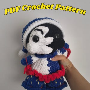 Puede incluir: Una muñeca de ganchillo con cara dividida en blanco y negro, ojos de seguridad negros, gorro azul y blanco, traje azul y blanco con volante rojo en la cintura y pantalones azules con detalles amarillos. El texto dice: PDF Crochet Pattern.