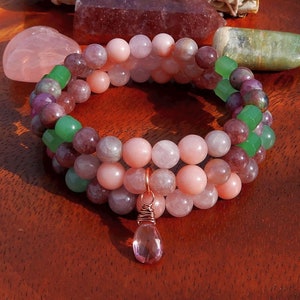 May include: A stack of three beaded bracelets with pink, purple, and green beads. A copper wire pendant with a pink crystal hangs from the bottom bracelet. The bracelets are on a wooden tray with crystals and sage.
