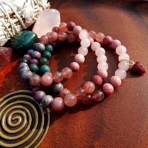 May include: Three beaded necklaces with varying shades of pink, red, and green gemstones. One necklace has a small red charm. Other items include a sage bundle, a rose quartz crystal, and a brass spiral on a wooden surface.