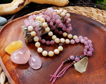 May include: A mala or prayer bead necklace made with pink and yellow gemstone beads. The necklace is on a wooden tray with a bundle of sage and three loose gemstones.