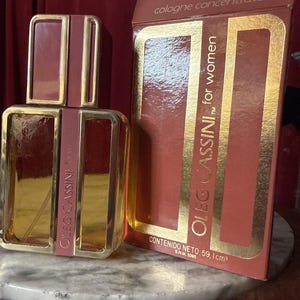 May include: A rectangular glass perfume bottle and its matching cardboard box, both featuring a deep reddish-brown color with metallic gold trim. The text on the box reads cologne concentrate, OLEG CASSINI for women, and CONTENIDO NETO: 59,1 cm³.