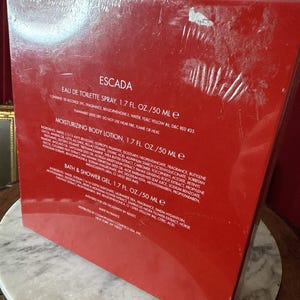May include: A red rectangular gift set box featuring the brand name ESCADA. The text lists an Eau de Toilette spray, a moisturizing body lotion, and a bath and shower gel, each containing 1.7 fl. oz. or 50 ml. The box includes ingredient lists.