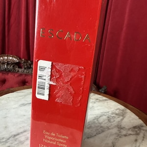 May include: A rectangular red perfume box with the brand name ESCADA printed in gold lettering at the top. The lower portion features the text Eau de Toilette Vaporisateur Natural Spray 1.7 FL. OZ./50 ml. A white barcode sticker is attached.